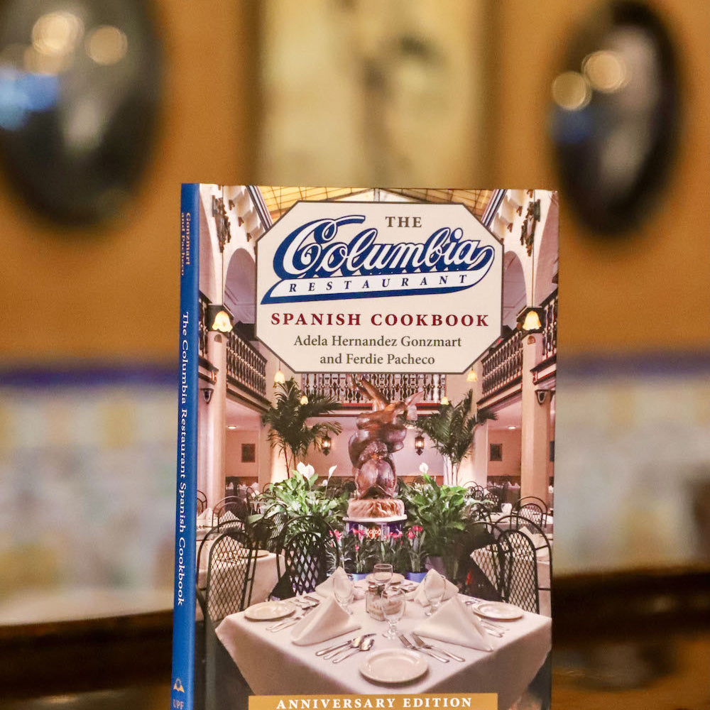 The image shows the cover of the 'Columbia Spanish Cookbook' with a foreword by Andrea Gonzmart Williams. The book is placed on a table with a restaurant setting in the background.