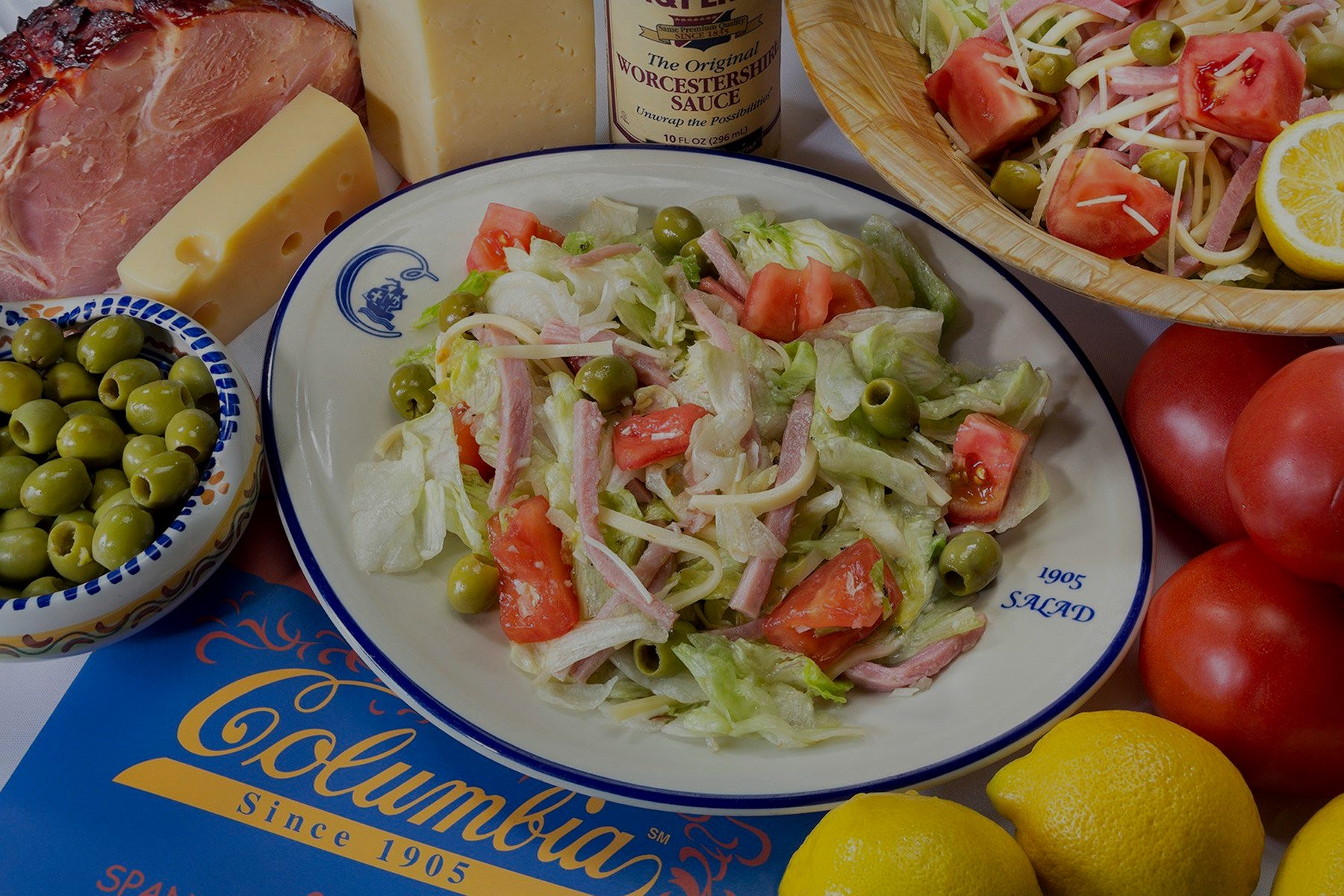 Salad with ham, cheese, and vegetables on a Columbia brand plate, surrounded by lemons, tomatoes, and a bottle of Worcestershire sauce.