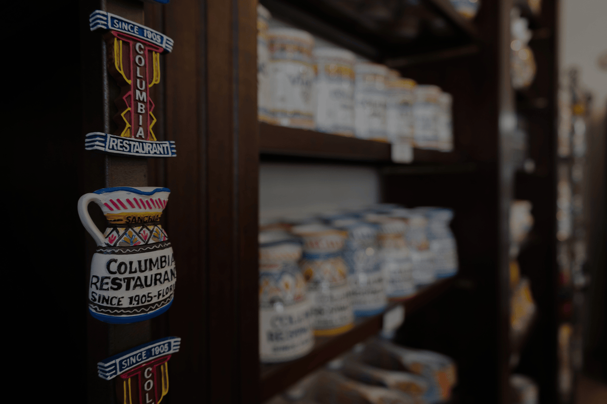 A close-up of a dark wooden shelf displaying colorful souvenir magnets or stickers featuring the 'Columbus Restaurant' logo, with blurred mugs on the shelving in the background.