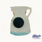 A ceramic pitcher with a textured off-white glaze, featuring a large black circular motif on the front, a light blue painted base, and small colorful paint splatters. The image is set against a plain white background with a 'Columbus Spanish Restaurants' logo in the bottom right corner.