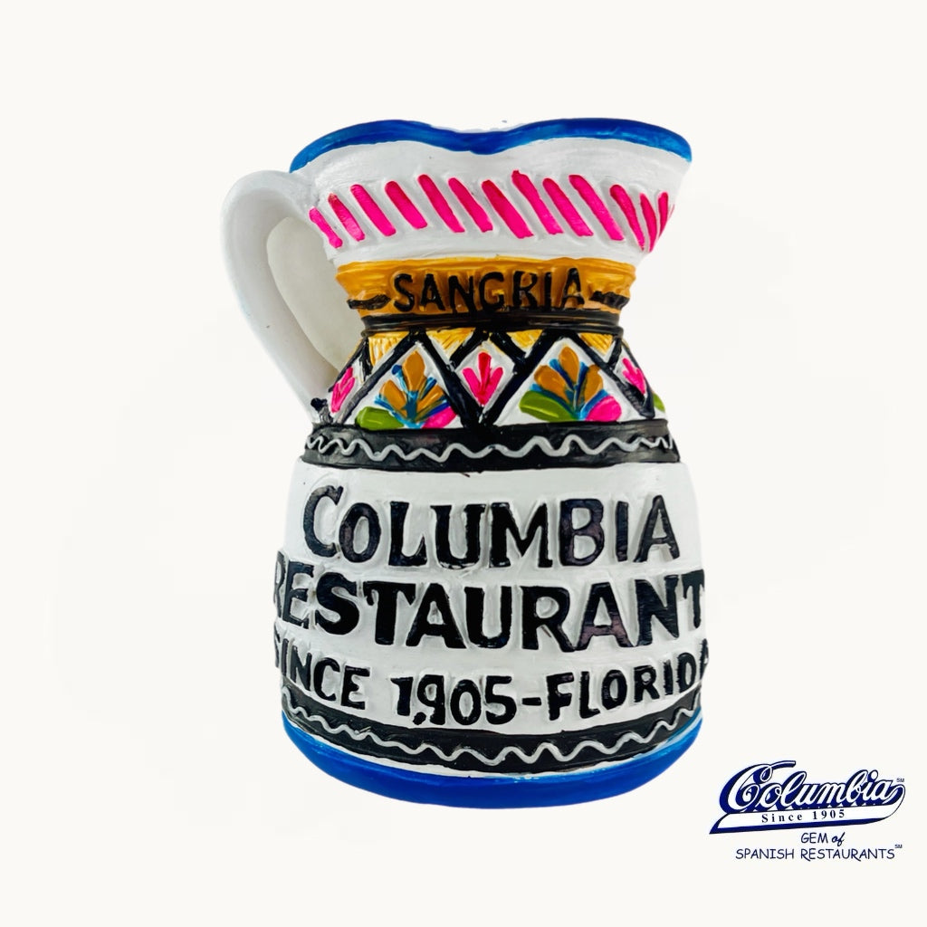 A ceramic pitcher styled as a vintage sign for 'Columbia Restaurant' in San Genclo, Florida, featuring a white body with colorful hand-painted details in blue, pink, yellow, and black, set against a plain white background.